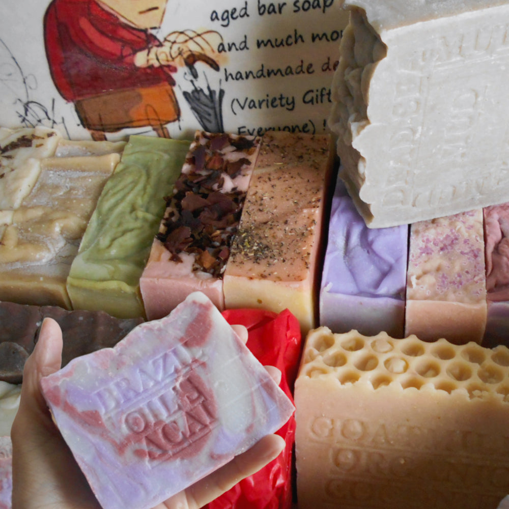 A hand holds a pale pink and red bar of soap embossed with 'BRAZI OIL & ALOHA' in the foreground, with multiple other colorful handmade soaps stacked behind it, including green, pink, purple, red, and beige varieties handmade soap 