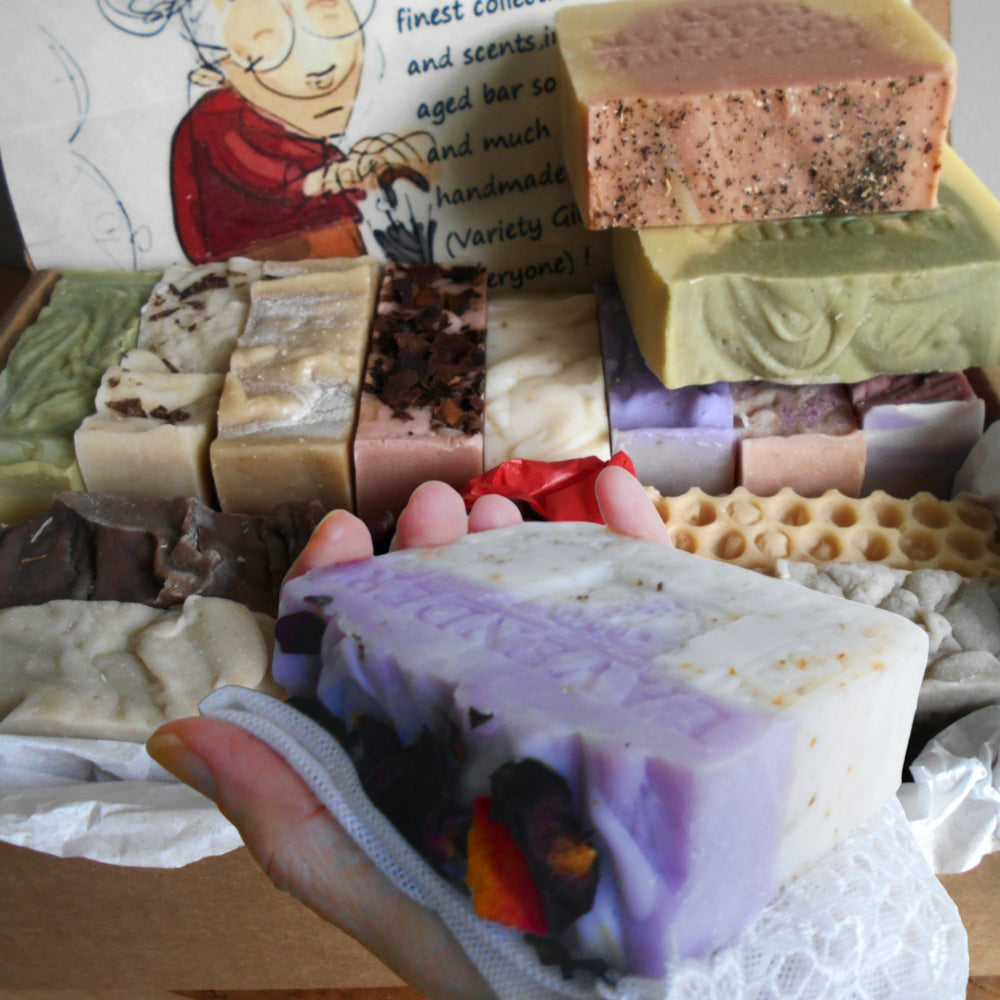 A hand holds a lavender and white layered soap bar in a box filled with multiple artisan soaps in various colors and textures, with a printed card in the background. All soaps handcrafted 