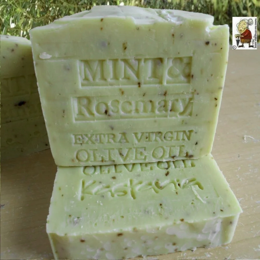 A handmade bar of soap labeled 'MINT & Rosemary EXTRA VIRGIN OLIVE OIL' stacked on top of another bar on a wooden surface, with a small cartoon logo in the top right corner.