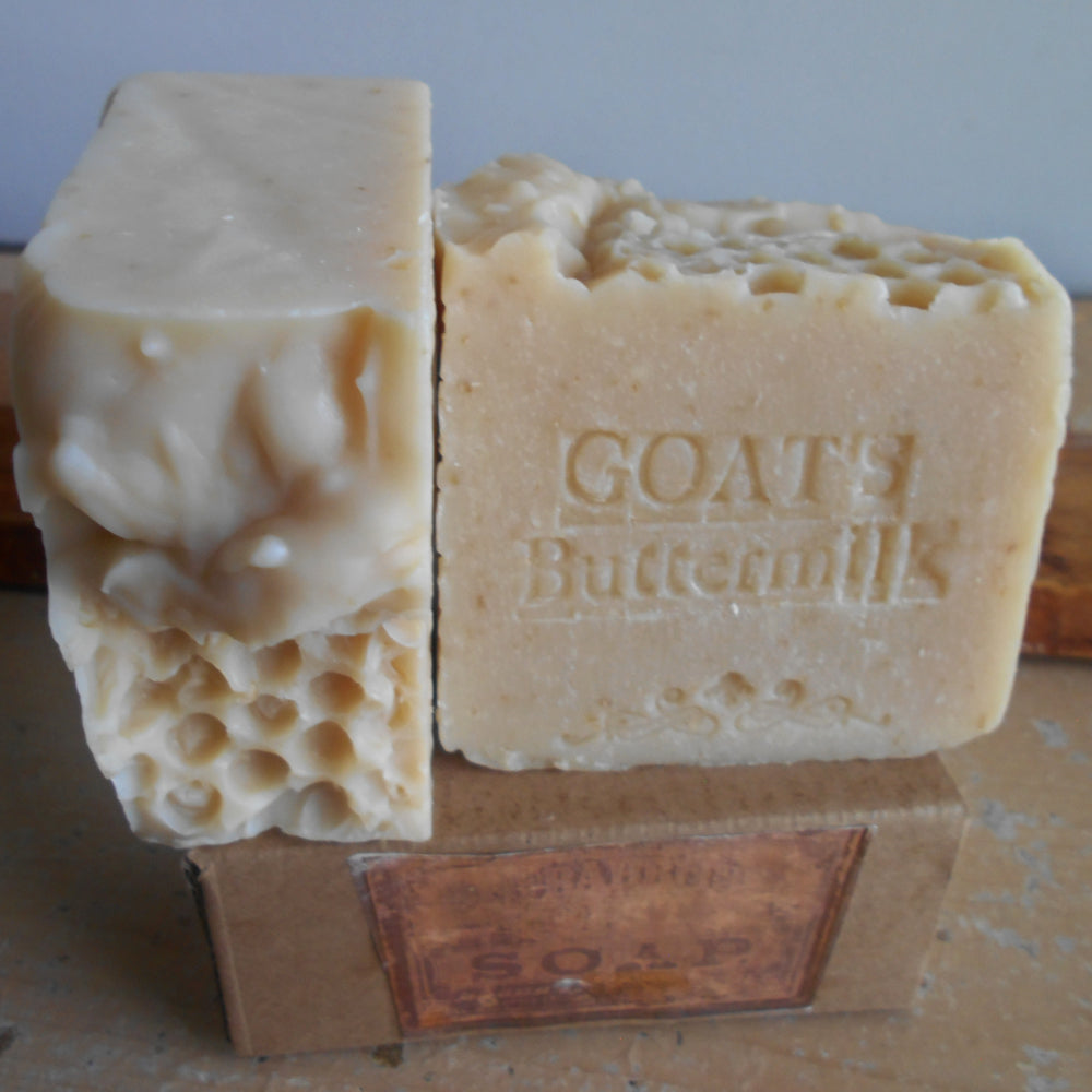  handmade bars of goat buttermilk soap standing upright on a cardboard box. One bar is shown from the side with a honeycomb cut pattern, and the other is shown from the front with the brand name 'GOAT'S Buttermilk' embossed on it.