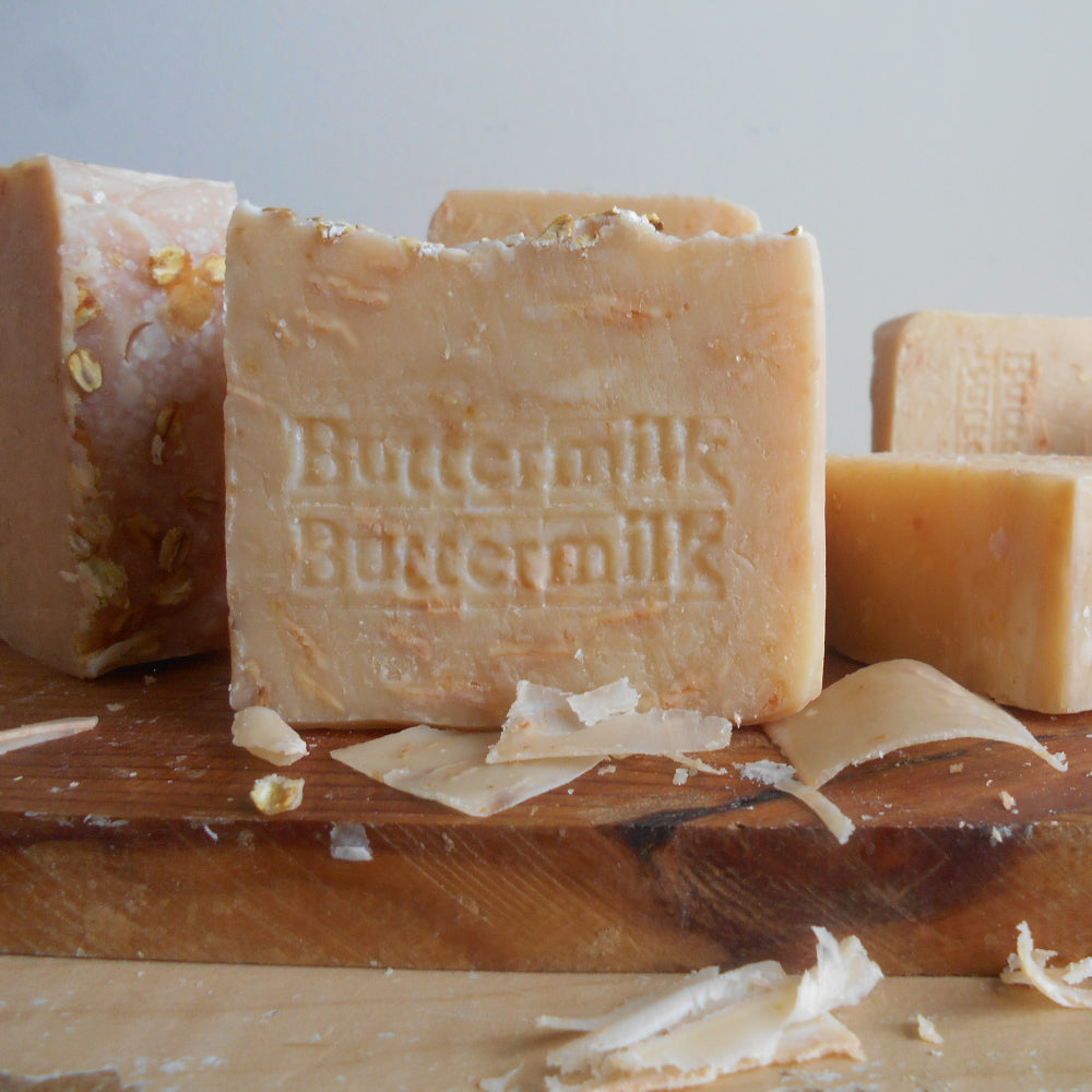 A close-up of handmade rectangular soap bars in a warm beige tone, with the center bar embossed with the words 'Buttermilk Buttermilk.' The soaps are arranged on a wooden board with shaving shavings scattered around, against a soft light gray background. 