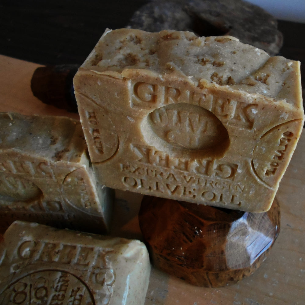 Close-up of two bars of Greek extra virgin olive oil soap, enhanced with Mediterranean Sea Salt. One bar is propped up and detailed with an engraved label. The soap bars have a natural, rustic appearance, with some texture on the surface, and are displayed on a wooden stand.