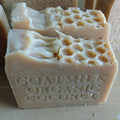 A close-up image of two bars of Grandma All Natural Soap, each with honeycomb-like indentations on top. The soap has "GOATMILK ORGANIC COCONUT" embossed on its side, indicating its ingredients sourced from local farms and never powdered or diluted with water. The bars are light beige and have an artisanal appearance.