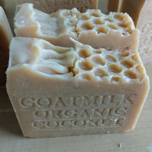A close-up image of two bars of Grandma All Natural Soap, each with honeycomb-like indentations on top. The soap has "GOATMILK ORGANIC COCONUT" embossed on its side, indicating its ingredients sourced from local farms and never powdered or diluted with water. The bars are light beige and have an artisanal appearance.
