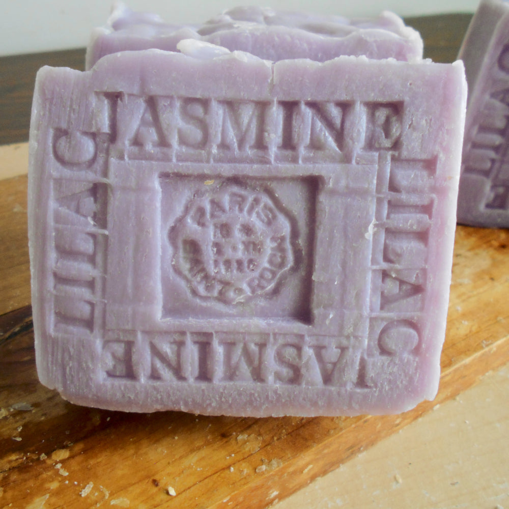 A detailed bar of French Jasmine Lilac Soap with Organic Shea Butter embossed with "Lilac" and "Jasmine" on its surface. The square-shaped bar, featuring a crown motif and the text “Paris” and “Paris 1823,” is purple with a textured, rustic appearance.