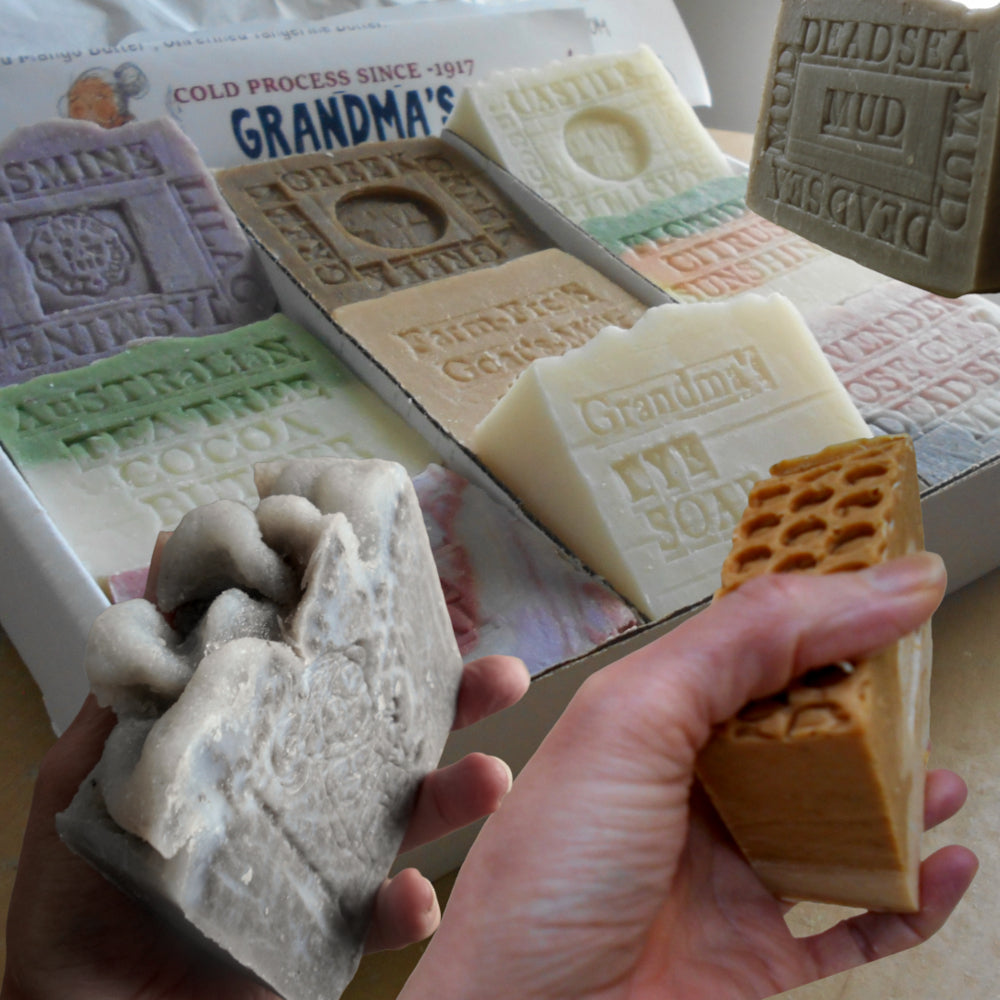 A close-up of hands holding handmade soap bars in front of a display of assorted soaps in a box. The soaps have rustic, textured designs and embossed bar names, including 'Grandma's', 'Dead Sea Mud', and 'Australian Cocoa  Jasmine bar soaps 