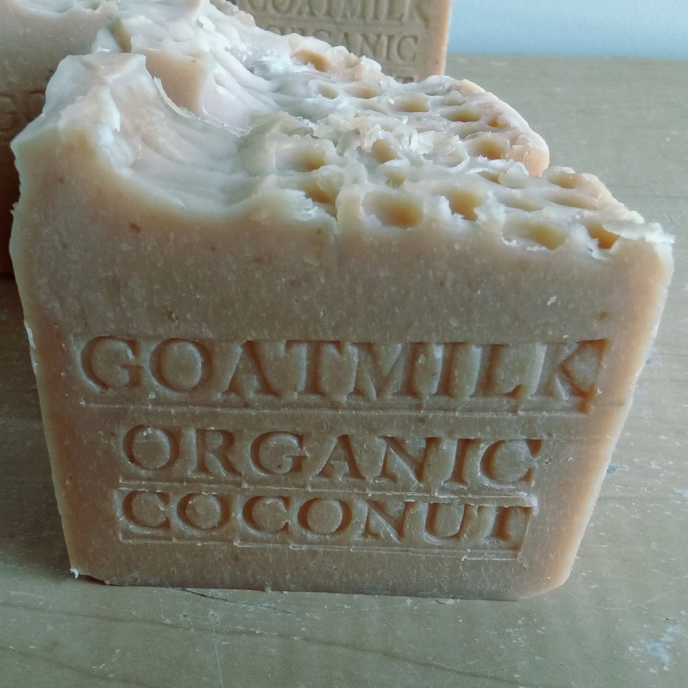 Two beige, rectangular bars of Goat Milk and Organic Coconut Milk Soap are engraved with the words "GRANDMAS ALL NATURAL SOAP." The soap appears textured, with slightly uneven edges and a honeycomb-like pattern on the top surface. Made from Goat Milk and Organic Coconut Milk Soap, it acts as a natural exfoliant. The background is a plain, light-colored surface.