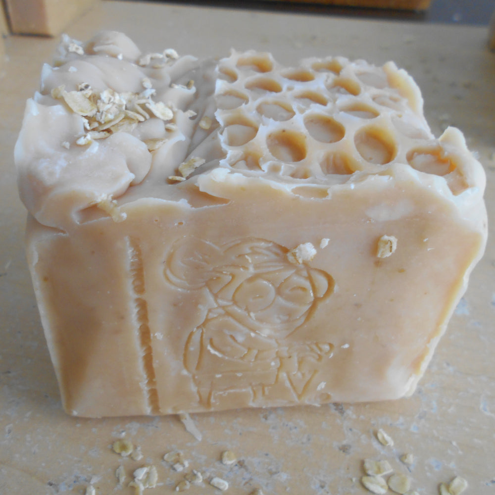 A handmade rectangular bar of soap in a light milk color, featuring a honeycomb pattern on one half, rolled oats on top, and an embossed  grandma's with a umbrella  on the back on the bar , set on a light wooden surface.