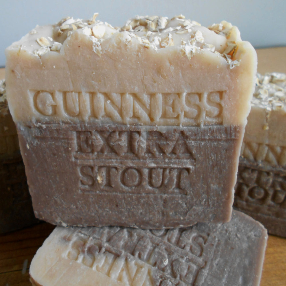 Bar of Guinness Extra Stout soap with visible branding on a wooden surface.  Two bars of handcrafted soap are shown. The bar in the front, made with Guinness beer soap, features layers of dark brown, light brown, and beige with "STOUT GUINNESS" embossed on it. The rough-textured soap includes oatmeal pieces embedded in the beige layer at the bottom and helps soften your skin.
