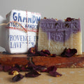 Two lavender soap bars displayed on a rustic wooden block with scattered dried rose petals. One soap bar is wrapped in white paper with blue text reading 'Grandm Natural Handcrafted Provence French Lavender,' and the other is unwrapped to show a lavender-tinted top and cream-colored bottom with 'Lavender' embossed on the front.