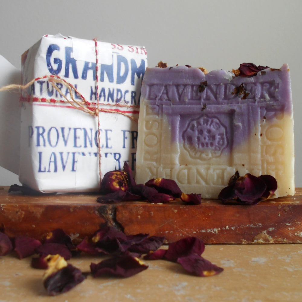 Two lavender soap bars displayed on a rustic wooden block with scattered dried rose petals. One soap bar is wrapped in white paper with blue text reading 'Grandm Natural Handcrafted Provence French Lavender,' and the other is unwrapped to show a lavender-tinted top and cream-colored bottom with 'Lavender' embossed on the front.