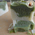 A bar of Grandmas All Natural Soap’s Australian Tea Tree Soap with Organic Spirulina, featuring green and white layers and soap shavings around it, sits on a wooden surface. A round logo appears in the upper right corner.