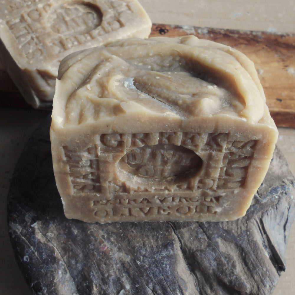 A Bar of Handcrafted soap  and Sea Salt  Greek olive oil   embossed lettering, resting on a dark textured stone slab with another bar in the back 
