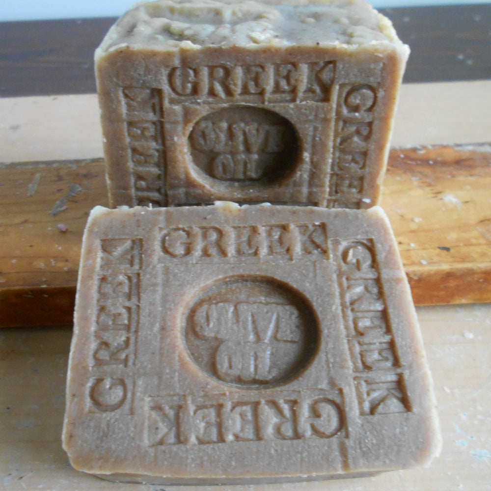 Two rectangular bars of Greek Olive Oil soap stacked on a wooden surface. The soap is a natural light brown color with embossed text reading 'GREEK' and 'OLIVE OIL'  Soap 