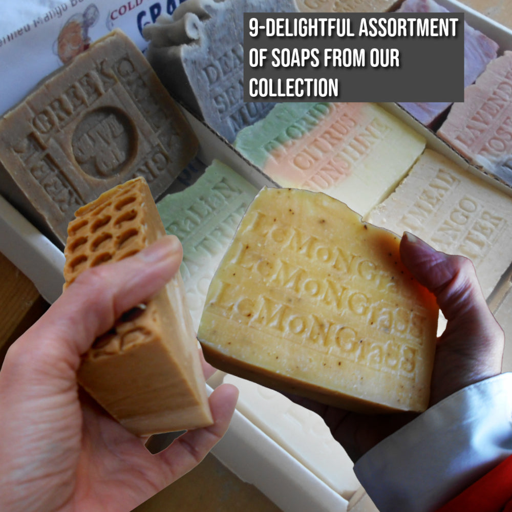 Two hands holding artisanal soaps in front of a tray of multiple soaps. One soap is a rectangular bar with a honeycomb pattern on one end, and the other is a large rounded bar embossed with 'Lemongrass