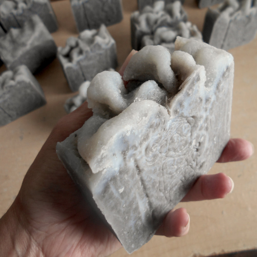 A hand holds a rectangular handmade dead sea mud soap bar with a highly textured, and a stamp of grandma's soap