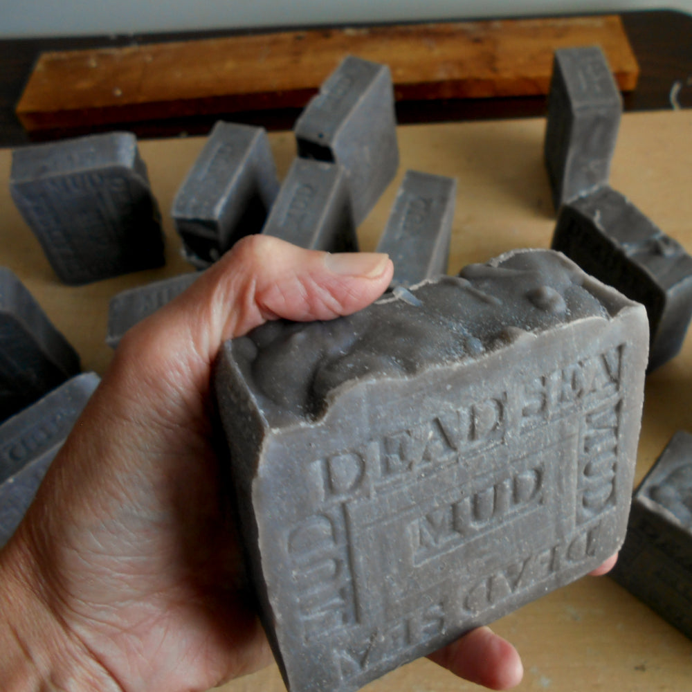 A hand holds a gray rectangular bar of soap embossed with  Dead Sea Mud in a workshop setting with multiple similar soap bars scattered in the background. Dead Sea Mud Soap - Google shop - Natural Mud Soap from The Dead Sea