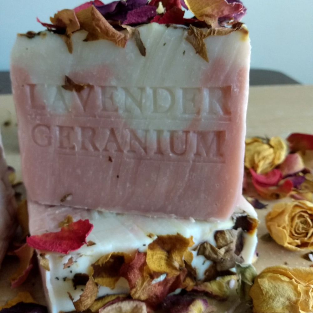 A close-up of a handmade soap bar labeled 'LAVENDER GERANIUM,' with dried rose petals and petals scattered around it on a neutral surface.