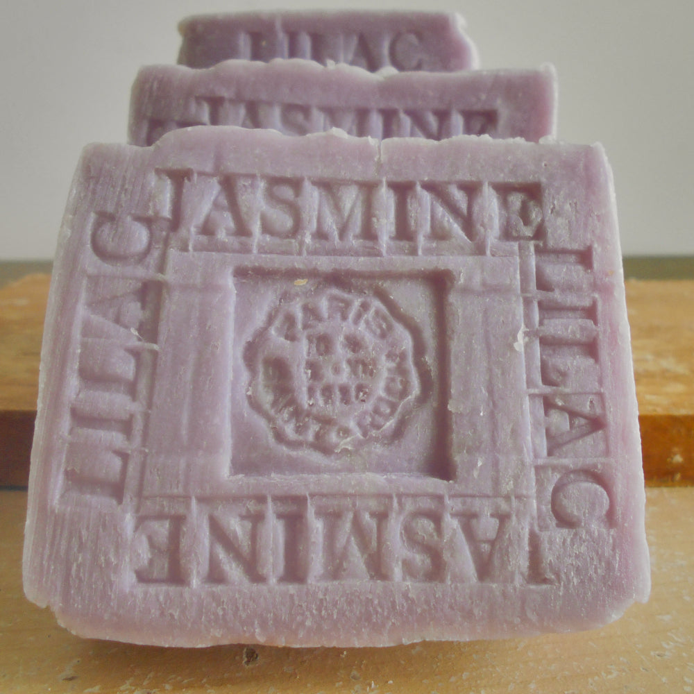 3 bars of lavender lilac soap stacked on a wooden surface, with the front bar embossed with 'JASMINE' and 'LILAC'.