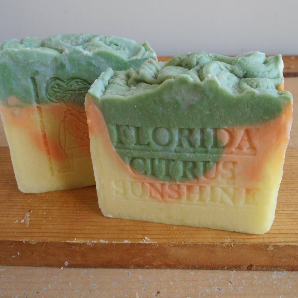 handmade soap bars with a green, orange, and pale yellow marbled design, one embossed with 'FLORIDA CURUS SUNSHINE', displayed on a wooden board.