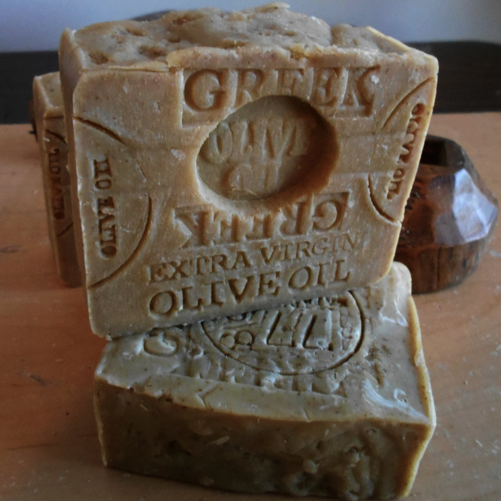Close-up of two bars of Greek extra virgin olive oil soap, enhanced with Mediterranean Sea Salt. One bar is propped up and detailed with an engraved label. The soap bars have a natural, rustic appearance, with some texture on the surface, and are displayed on a wooden stand.