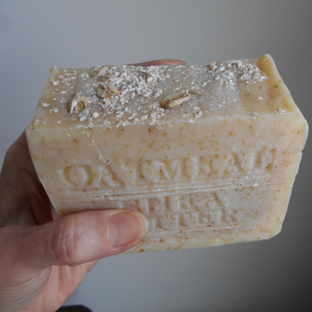 Bar of soap with visible branding held by a hand against a neutral background
textured bars of Organic Oatmeal Soap with Organic Shea Butter from Grandmas All Natural Soap is embossed with "OATMEAL SHEA BUTTER." Made with natural ingredients, the soap features visible specks 
