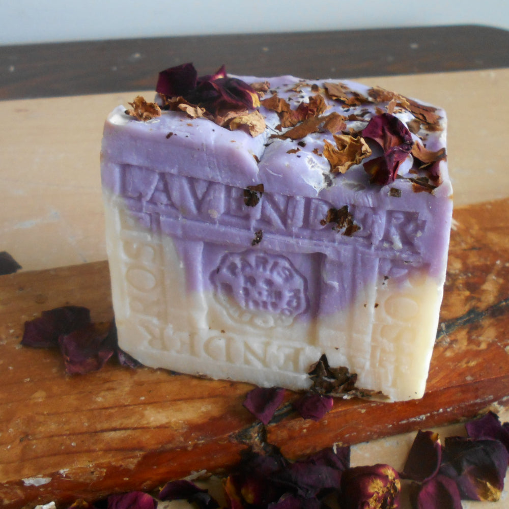 A rectangular bar of lavender soap with dried rose petals on top, resting on a rustic wooden board with scattered petals around it.