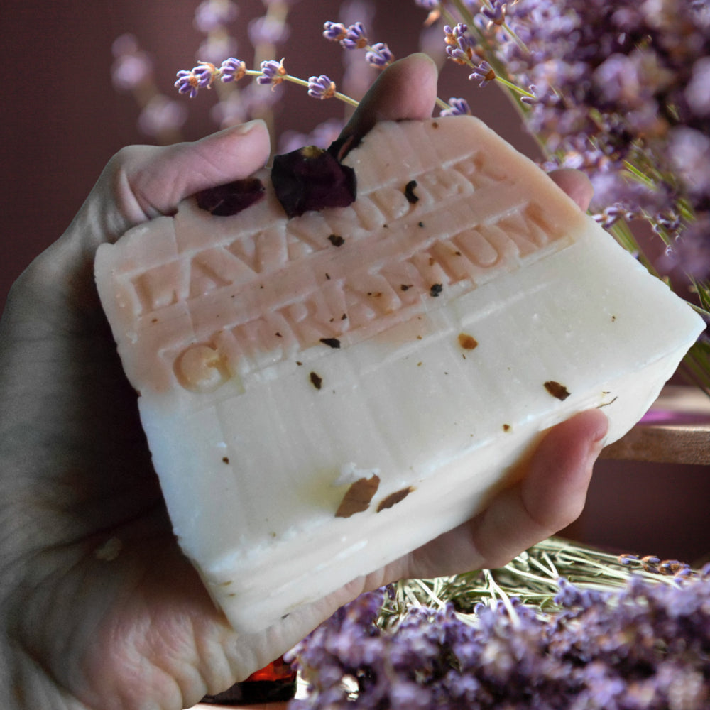 A hand holds a thick bar of lavender-geranium infused soap. The soap is two-toned, with a pale pink top half embossed with the text 'LAVENDER GERANIUM' and a white bottom half, with dried lavender buds and brown botanical inclusions embedded in it.