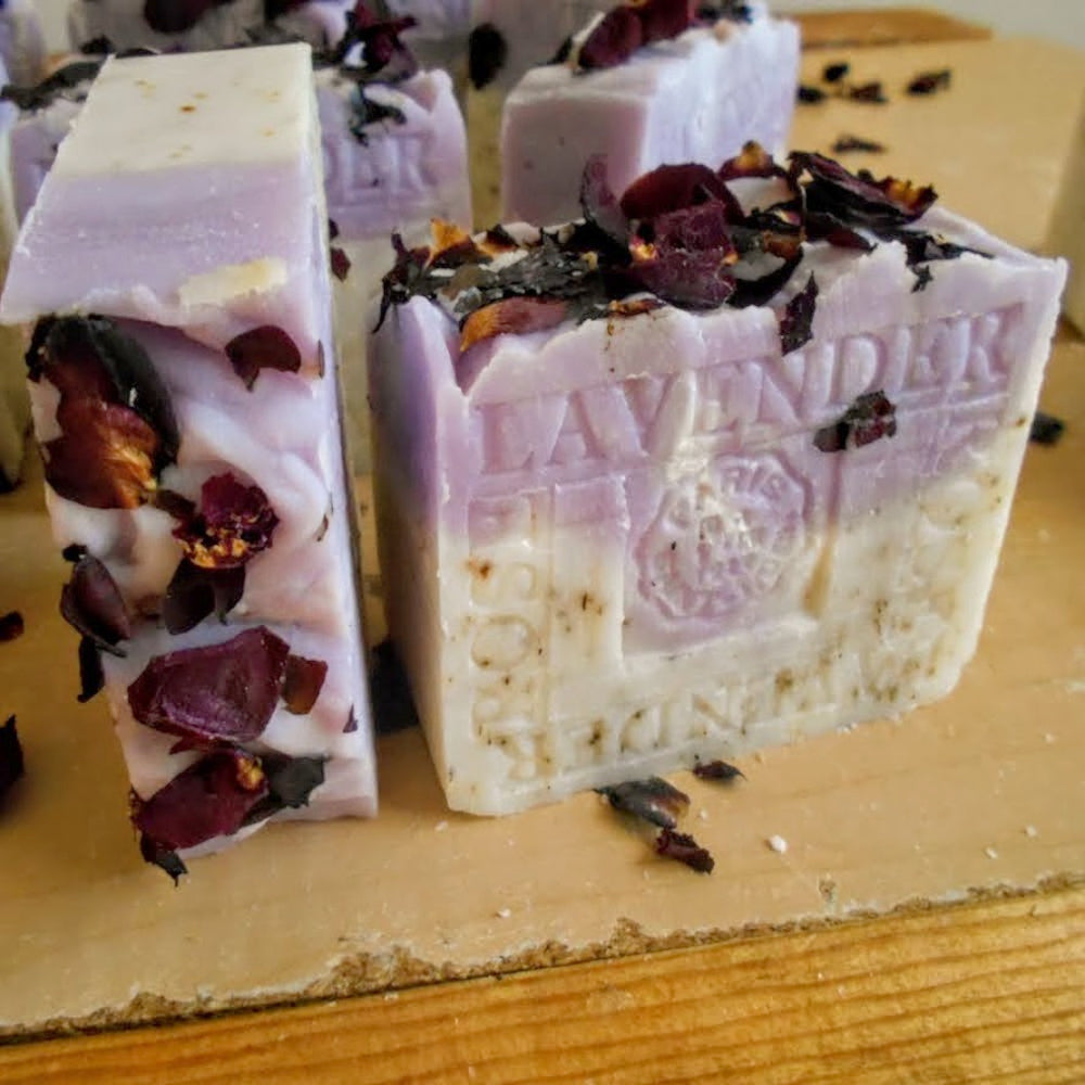 Handmade lavender soap bars with dried rose petals, displayed on a wooden board. One bar is standing upright on the left, and two more are partially visible in the background.