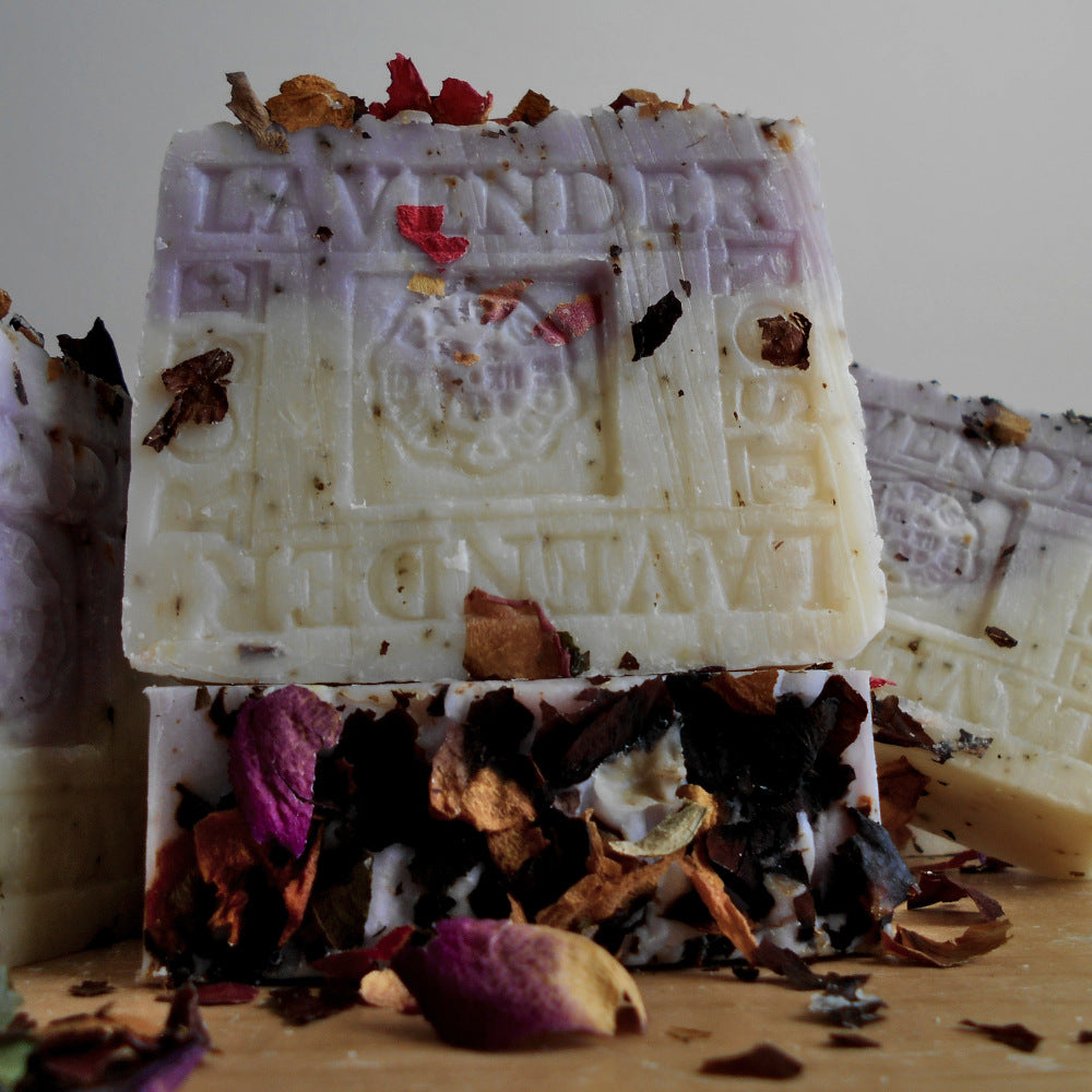 Close-up of several Provence French Lavender Handmade All Natural Soap with Crushed Rose Petals bars by Grandmas All Natural Soap with embossed designs. The handmade soaps are decorated with scattered crushed rose petals in various colors, creating a natural, rustic appearance. The background is a neutral, soft focus, enhancing the soap’s details.