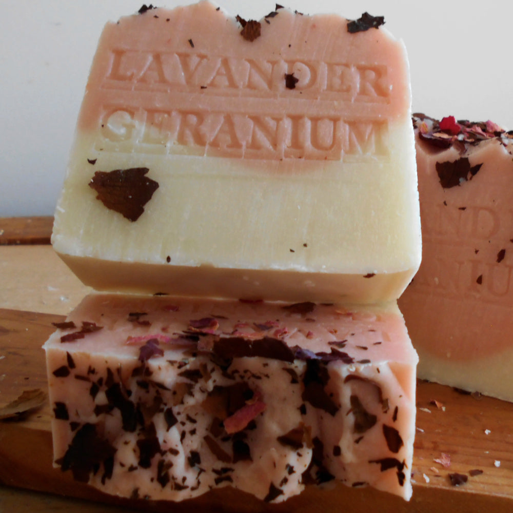 Lavender Geranium scented soap bars stacked on a wooden board, showing a pale pink and creamy yellow top layer with dark dried flower petals, and a cut bar revealing a pink base with embedded red petals.