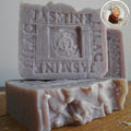 Three bars of Grandmas All Natural Soap’s French Jasmine Lilac Soap with Organic Shea Butter are stacked on a wooden surface. The top bar is stamped "JASMINE LILAC." A round logo with a person appears in the top right corner.