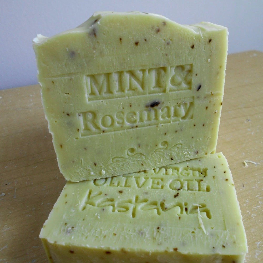 Two handmade bars of soap stacked on a wooden surface. The top bar is labeled 'MINT & Rosemary' and the bottom bar is labeled 'OLIVE OIL and  Rosemary Herbs