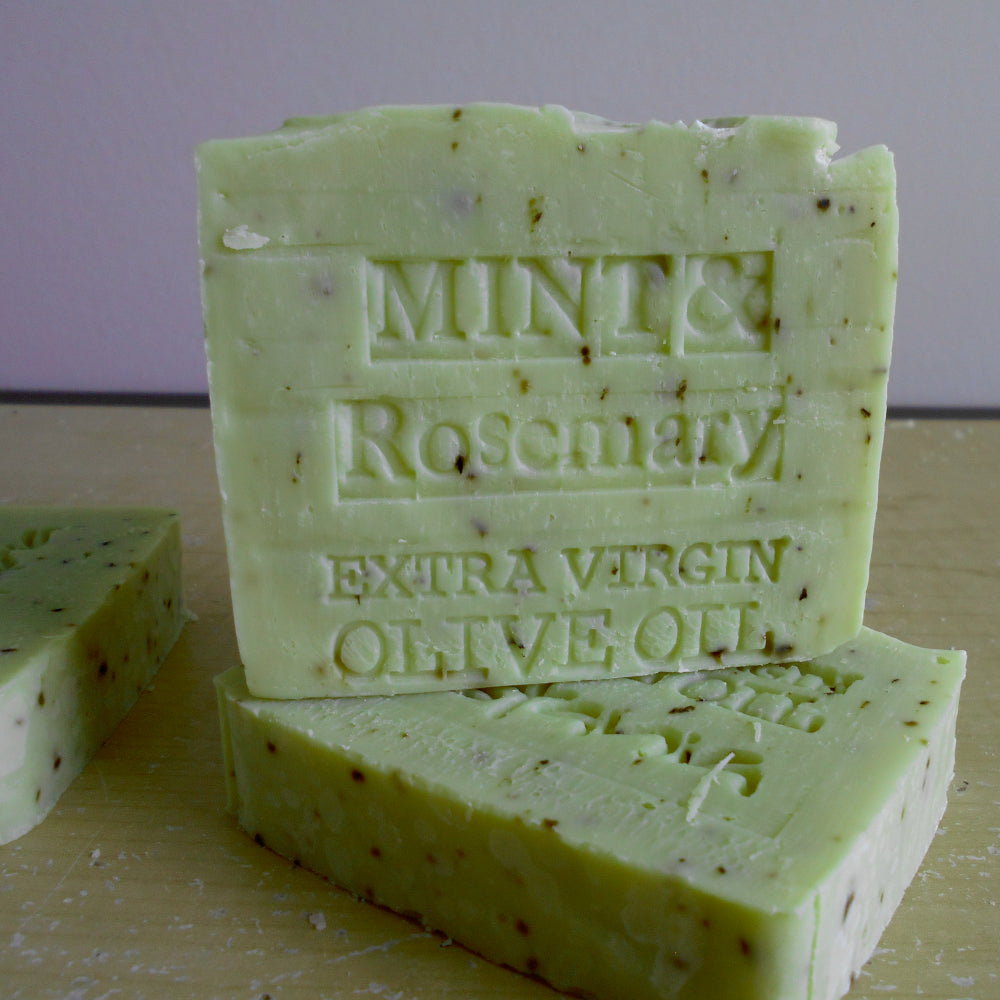 Three handmade bars of soap stacked on a light wooden surface, with the top bar embossed with 'MINT & Rosemary EXTRA VIRGIN OLIVE OIL'. The soap is a pale mint green with visible dark brown specks of herbs.
