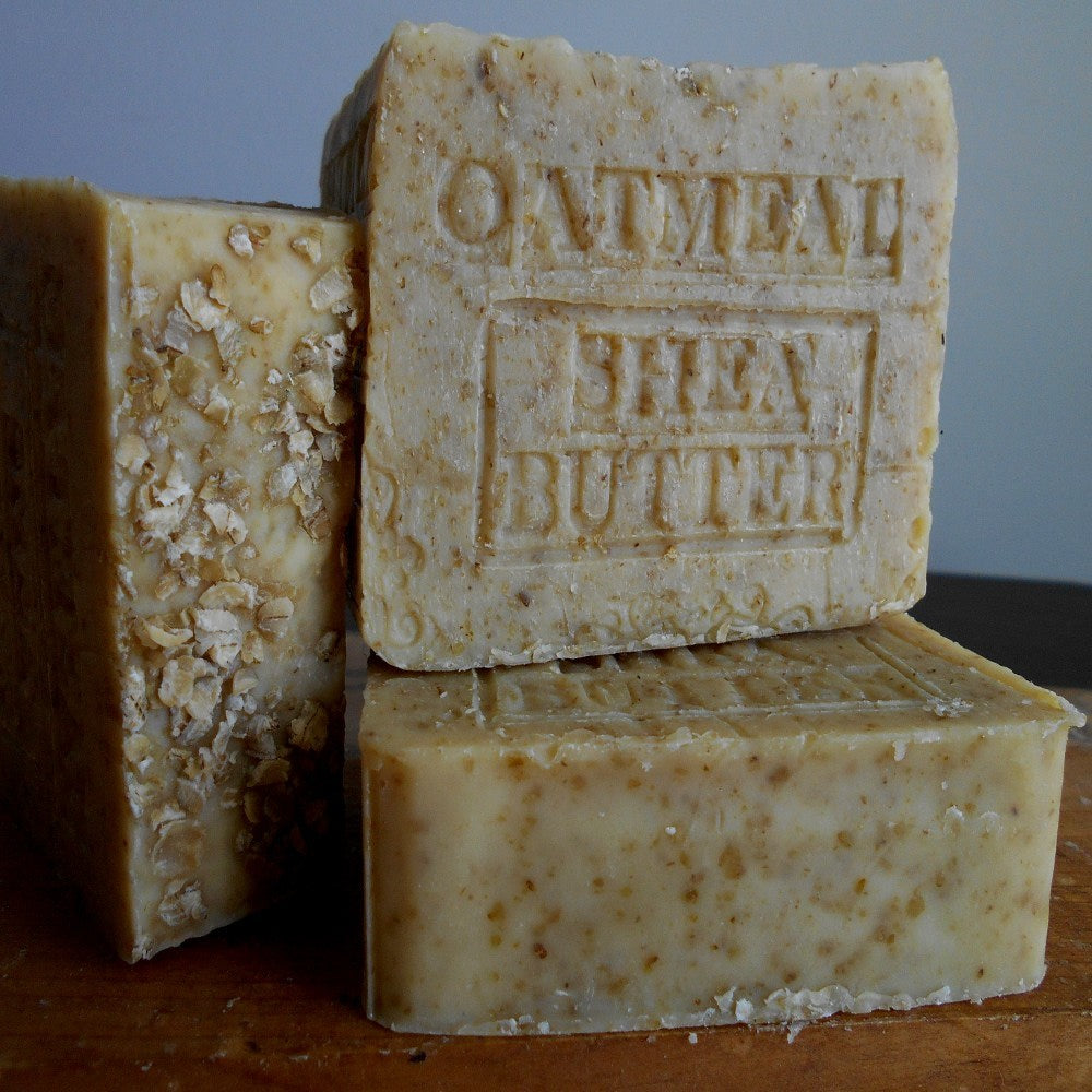 Three bars of oatmeal shea butter soap stacked on a wooden surface with a neutral background.