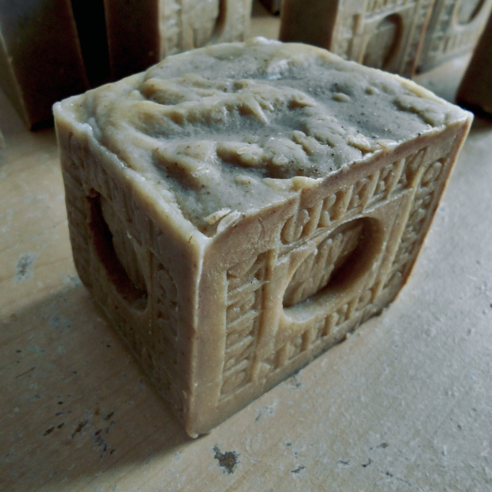 A handmade olive color cube-shaped bar of soap with embossed architectural details and the word 'GREEK Oil ' on the side, resting on a textured .