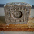  A Greek Olive Oil soap standing upright on a wooden surface. The soap is a natural  olive  color with embossed text reading 'GREEK OLIVE OIL'