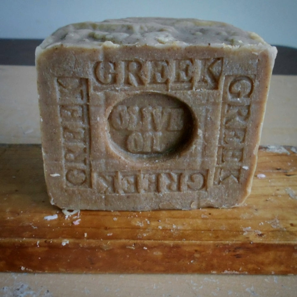  A Greek Olive Oil soap standing upright on a wooden surface. The soap is a natural  olive  color with embossed text reading 'GREEK OLIVE OIL'
