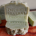 A handmade soap bar labeled 'Peppermint SEA CLAY Peppermint' in a pale greenish-gray color, standing upright on a reddish-brown surface with other soap bars blurred in the background.