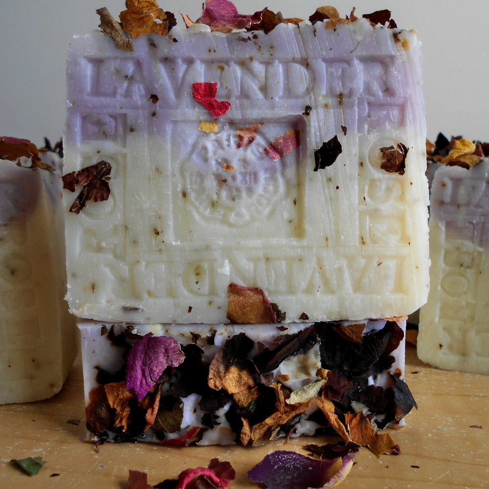 Handmade natural soap bars infused with Provence French lavender, featuring pieces of dried flowers embedded on the surface. The soap is a mix of white and light purple, with the word "LAVENDER" visibly embossed on each bar.  The soap bars are positioned on a wooden surface.