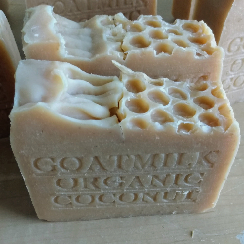  A close-up image of two bars of Grandma All Natural Soap, each with honeycomb-like indentations on top. The soap has "GOATMILK ORGANIC COCONUT" embossed on its side, indicating its ingredients sourced from local farms and never powdered or diluted with water. The bars are light beige and have an artisanal appearance.