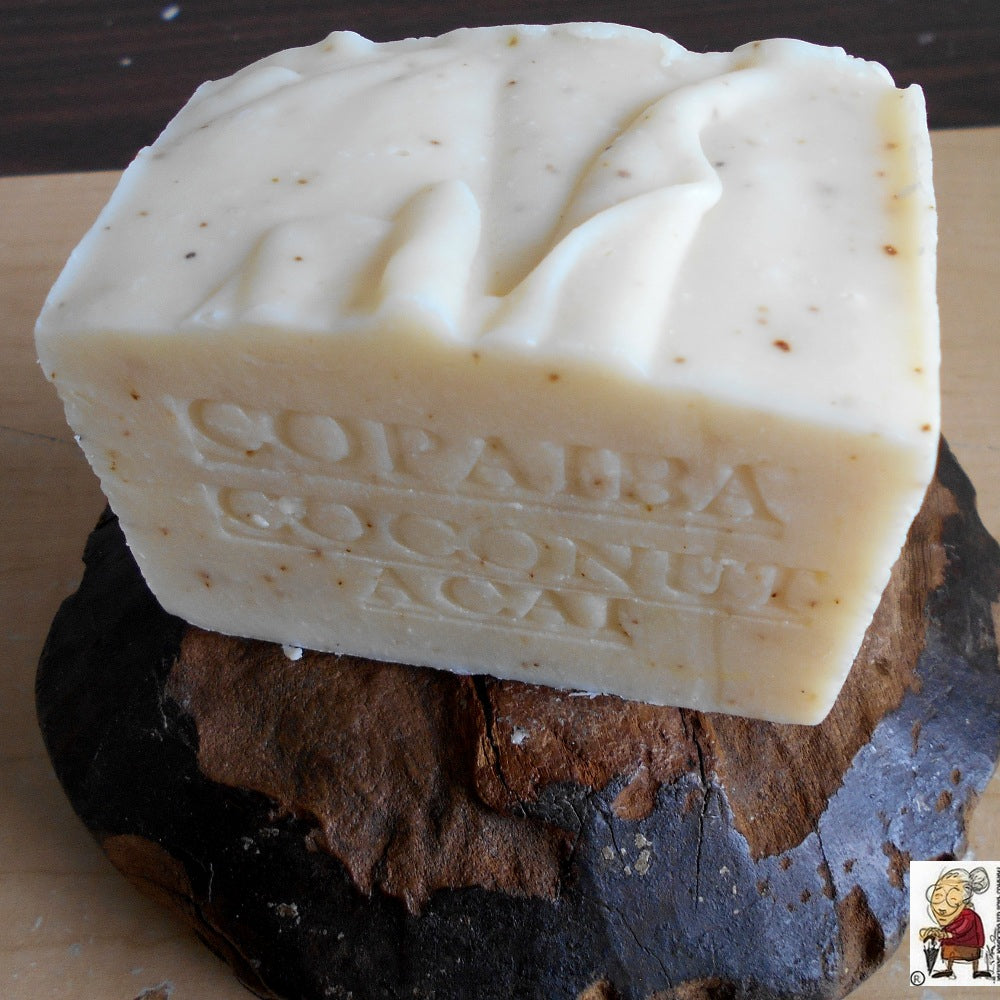  A rectangular beige soap bar with embossed text reading "Copaiba Coconut Açaí" resting on a dark brown stone. In the bottom right corner, there's a small logo featuring an illustration of a Grandmother. The soap has a textured surface with some speckles.