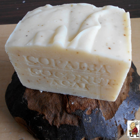  A rectangular beige soap bar with embossed text reading "Copaiba Coconut Açaí" resting on a dark brown stone. In the bottom right corner, there's a small logo featuring an illustration of a Grandmother. The soap has a textured surface with some speckles.