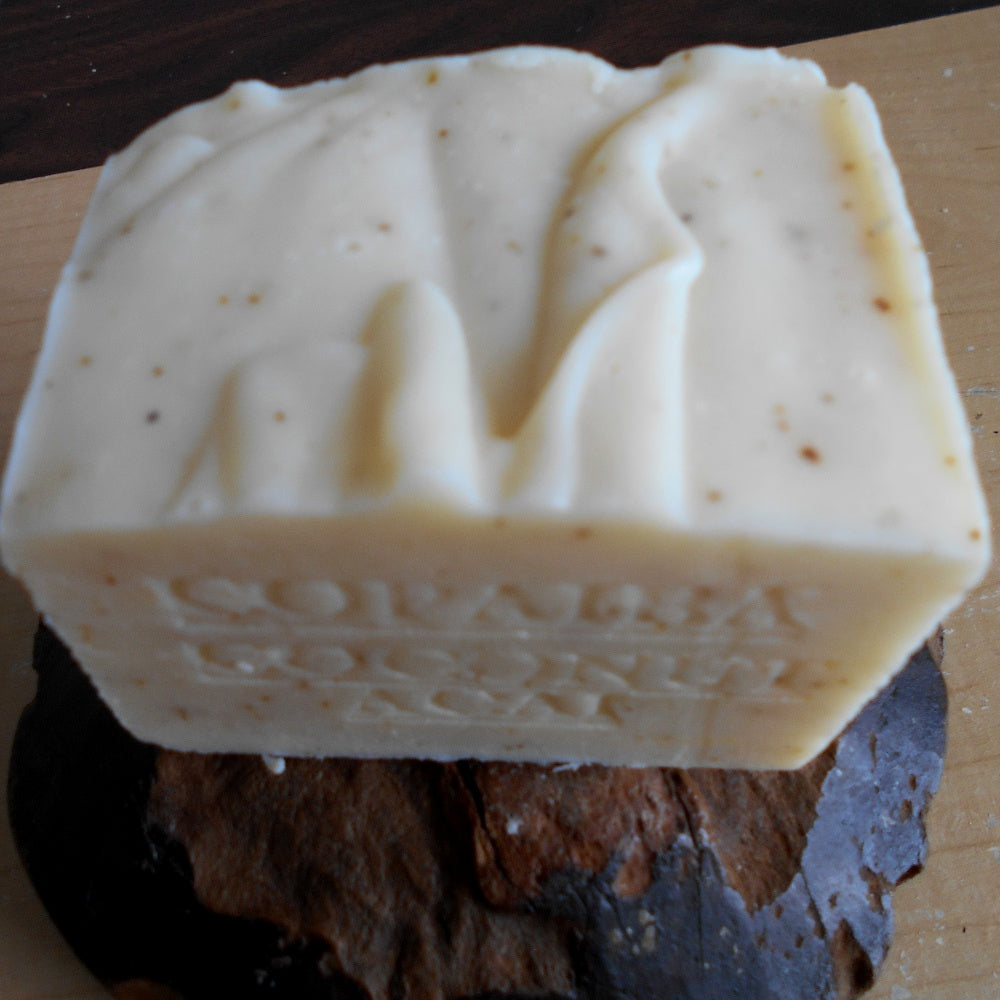 A close-up image of a rectangular, artisanal bar of soap with a creamy off-white color and small brown speckles. Infused with Organic Coconut Milk, the Aged Rainforest Copaiba with Coconut Milk Soap 12 OZ. by Grandmas All Natural Soap has an imperfect, handmade texture with indented lettering on the side. It is placed on a dark brown surface.