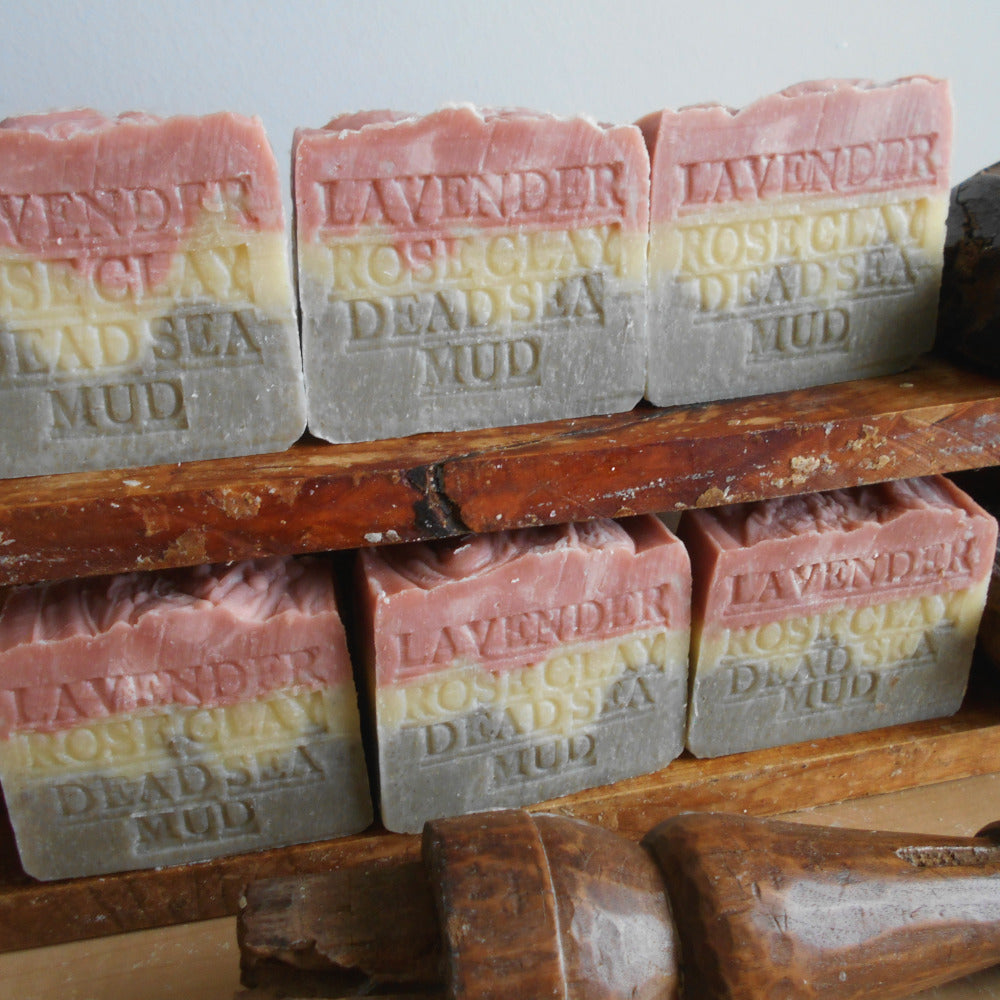 Provence Lavender Soap with Dead Sea Mud, French Rose Clay, and Shea Butter by Grandmas All Natural Soap features pink, cream, and gray layered bars stamped with the blend’s name, stacked neatly on wooden shelves.