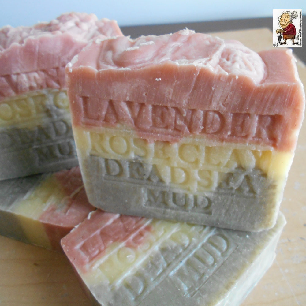 Three Provence Lavender Soap bars by Grandmas All Natural Soap—with Dead Sea Mud, French Rose Clay, and Shea Butter—feature pink, yellow, and gray layers embossed with “LAVENDER ROSE CLAY DEAD SEA MUD,” resting on wood. Brand logo top right.