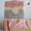 Two handmade soap bars are stacked; the top is Grandmas All Natural Soap's Provence Lavender Soap with Dead Sea Mud and French Rose Clay And Shea Butter, engraved on its surface. The bottom bar is pink. A small cartoon appears in the top right corner.