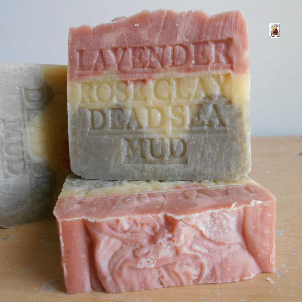 Two handcrafted bars from Grandmas All Natural Soap stacked on wood. The top, Provence Lavender Soap with Dead Sea Mud, French Rose Clay, and Shea Butter, is engraved “LAVENDER ROSE CLAY DEAD SEA MUD.” Layers of pink, yellow, and gray are visible.