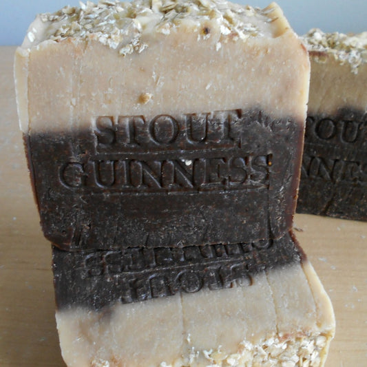  Two bars of handcrafted soap are shown. The bar in the front, made with Guinness beer soap, features layers of dark brown, light brown, and beige with "STOUT GUINNESS" embossed on it. The rough-textured soap includes oatmeal pieces embedded in the beige layer at the bottom and helps soften your skin.