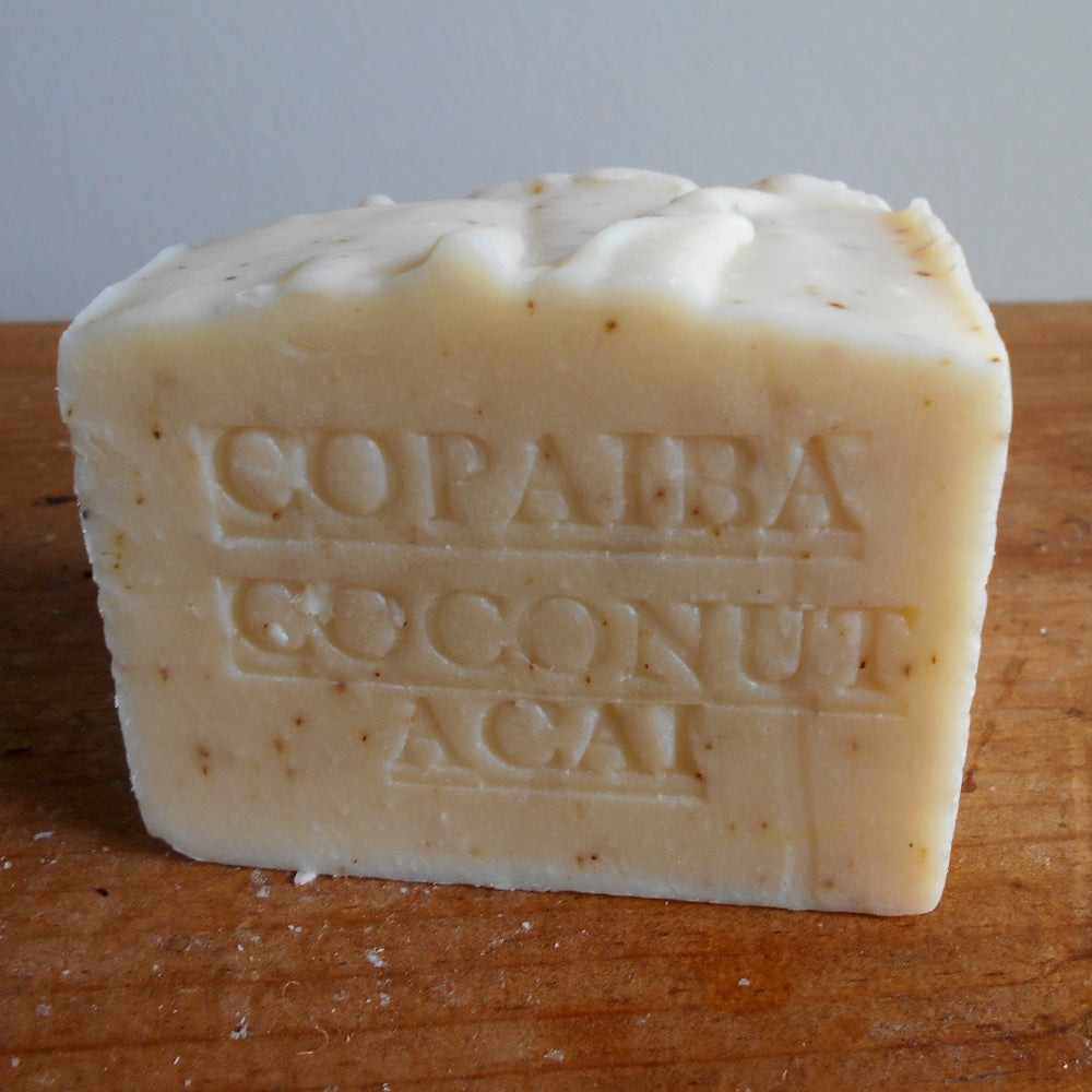 A bar of handcrafted soap labeled *Grandmas All Natural Soap: Aged Rainforest Copaiba with Coconut Milk Soap 12 OZ.* is placed on a wooden surface. The soap, infused with Copaiba Oil and Organic Coconut Milk, is off-white with some small brown specks. The texture appears slightly uneven, indicating it may be homemade or artisanal.